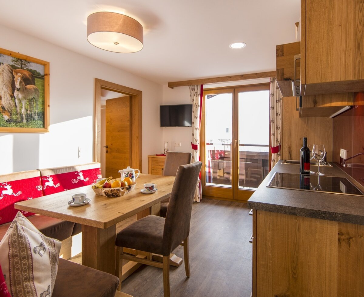 Der offene Wohnbereich des Ferienbauernhofs umfasst eine Küchenzeile, einen Essbereich mit Holztisch und Eckbank, einen Fernseher und Zugang zum Balkon.