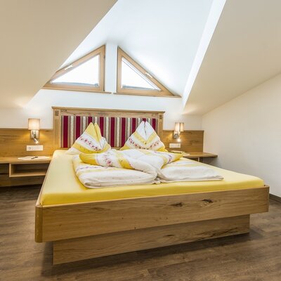 Doppelzimmer im Ferienbauernhof mit Holzbett, gepolstertem Kopfteil, Nachttischen und dreieckigen Dachfenstern.