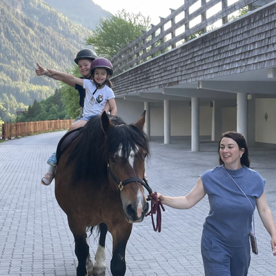 Kinder genießen einen Pferderitt, geführt von einem Erwachsenen, mit Bergen im Hintergrund.