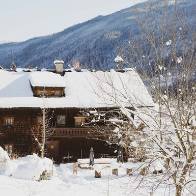 Das Ferienhaus im Winter, mit schneebedecktem Dach, Holzbalkon und einem Außensitzbereich. Copyright: Urlaub am Bauernhof / Pascal Baronit
