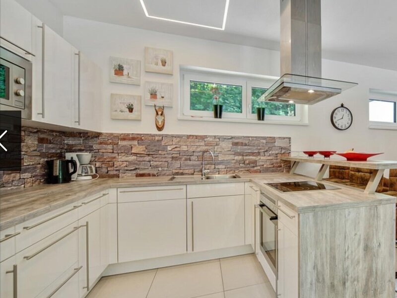 Moderne Küche der Ferienwohnung mit weißen Schränken, Steinoptik-Rückwand, Spüle, Herd, Backofen, Mikrowelle und Frühstückstheke.