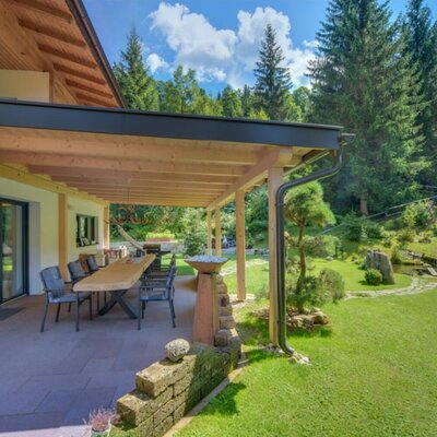 Überdachte Terrasse der Ferienwohnung mit großem Esstisch und direktem Zugang zum Garten mit Rasenfläche und Teich.