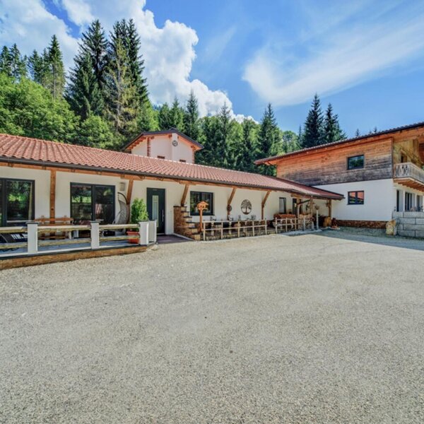 Außenansicht der Ferienwohnung mit großem Kiesparkplatz und einer Terrasse mit Sitzgelegenheiten, umgeben von Wald und Bergen.
