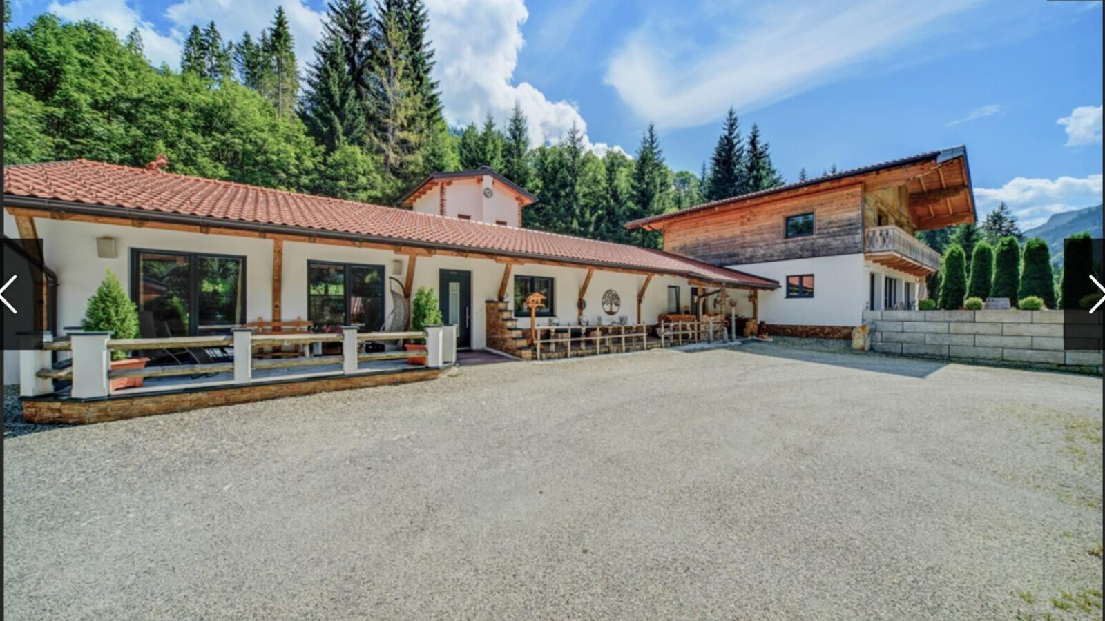 Außenansicht der Ferienwohnung mit großem Kiesparkplatz und einer Terrasse mit Sitzgelegenheiten, umgeben von Wald und Bergen.