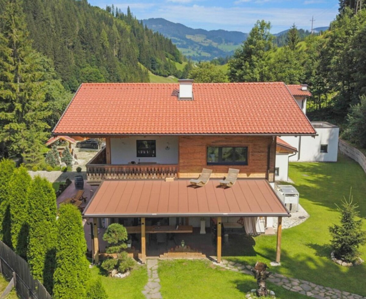 Außenansicht der Ferienwohnung mit rotem Ziegeldach, Holzbalkon, überdachter Terrasse und Garten, umgeben von bewaldeten Bergen.