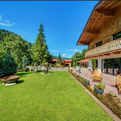 Außenbereich der Ferienwohnung mit großem Garten, Terrasse mit Liegestühlen und einer Feuerschale, umgeben von bewaldeten Bergen.
