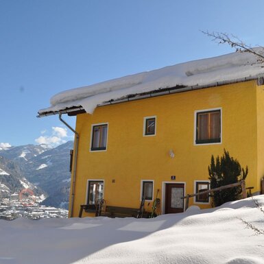 Der gelbe Hof im Winter, umgeben von Schnee, mit Skiausrüstung vor dem Eingang und Blick auf die umliegenden Berge und das Tal.