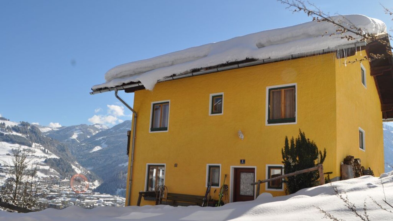 Der gelbe Hof im Winter, umgeben von Schnee, mit Skiausrüstung vor dem Eingang und Blick auf die umliegenden Berge und das Tal.