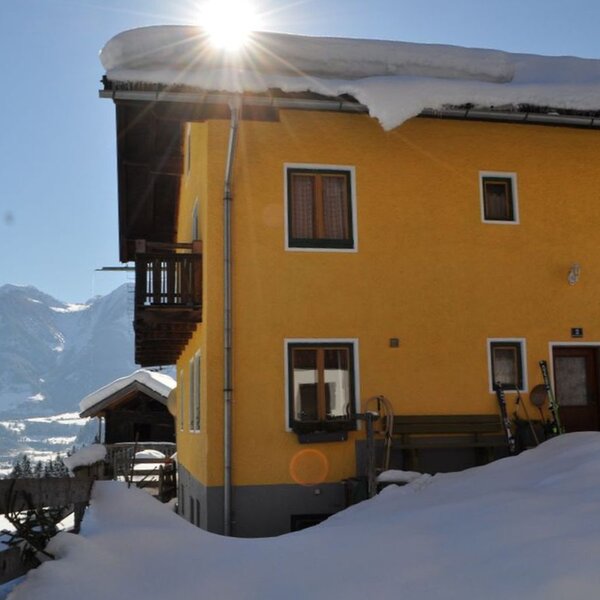 Der Hof im Winter mit gelber Fassade, schneebedecktem Dach, Skiern am Eingang und Bergblick.