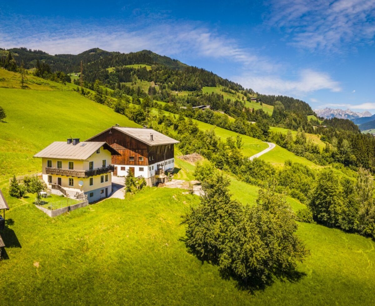Der Hof mit seinen Gebäuden, eingebettet in grüne Wiesen und umgeben von Bergen und Wäldern, bietet eine weite Aussicht auf das Tal.
