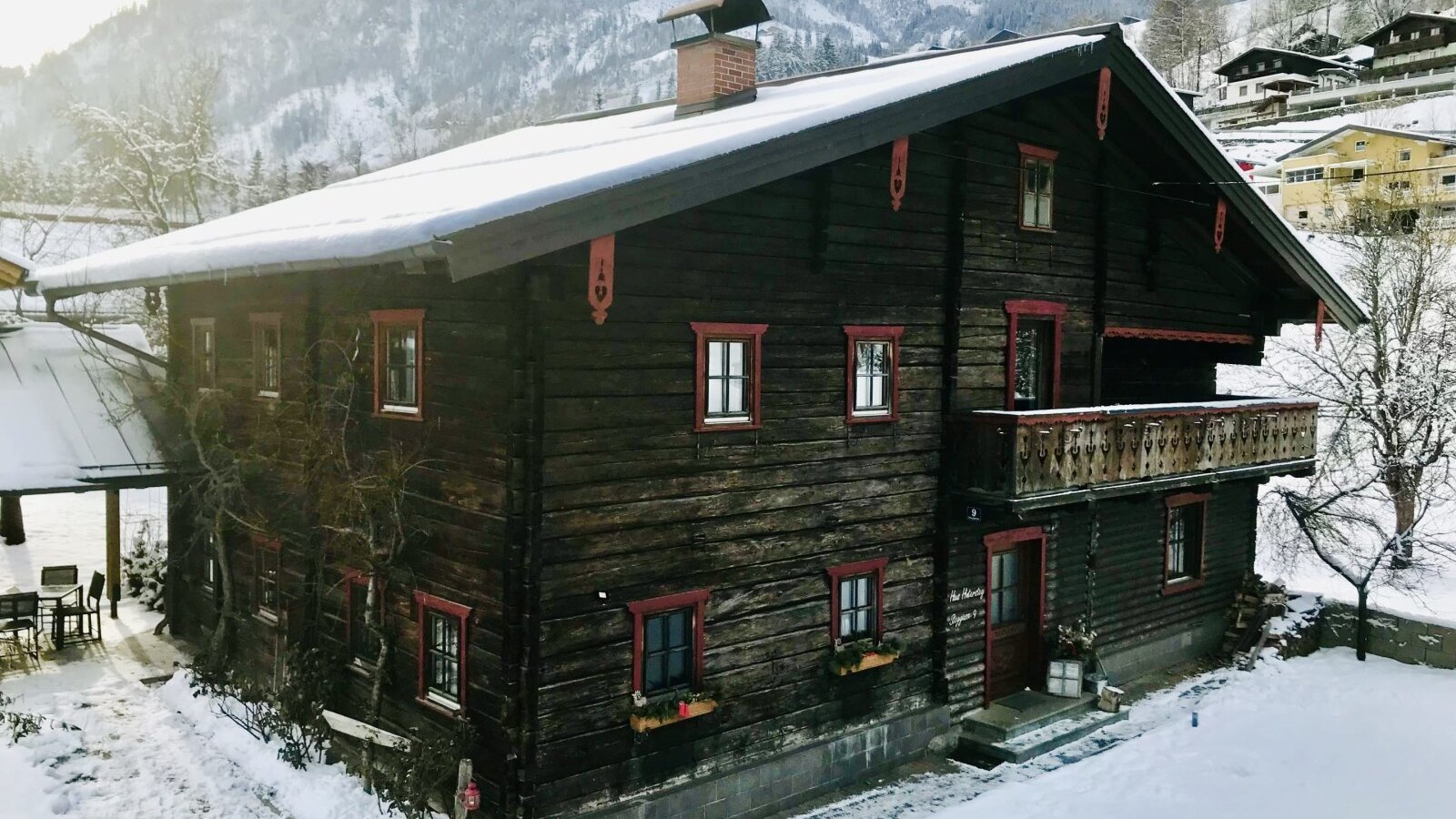 Der traditionelle Hof mit dunkler Holzfassade, Balkon und überdachtem Sitzbereich, umgeben von Schnee und Bergen.