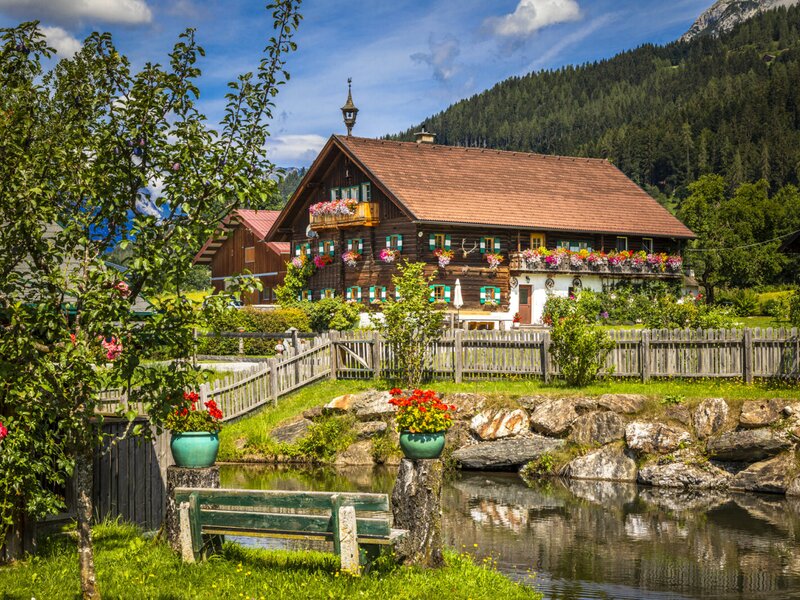 Der Hof mit dem traditionellen Bauernhaus, blumengeschmückten Balkonen, einem kleinen Teich und einer Sitzbank im Freien.