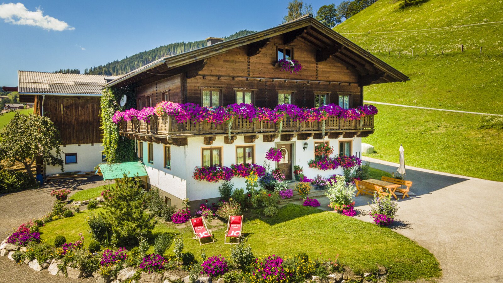 Der Bauernhof mit traditioneller Holzfassade, blühenden Balkonen, einem Vorgarten mit Liegestühlen und einem Sitzbereich im Freien.