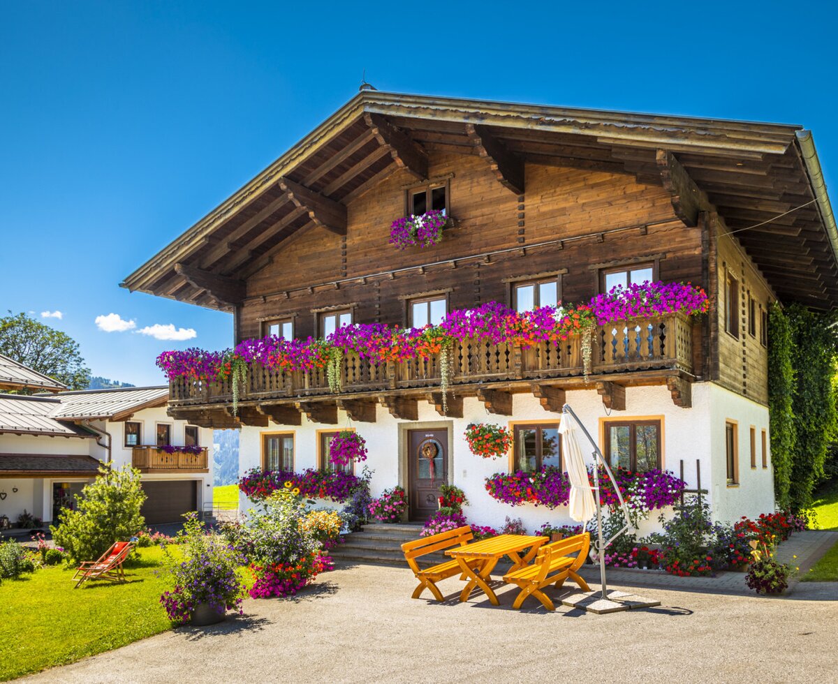 Der Bauernhof mit seiner traditionellen Holz- und Putzfassade, blumengeschmückten Balkonen und einem Sitzbereich im Freien.