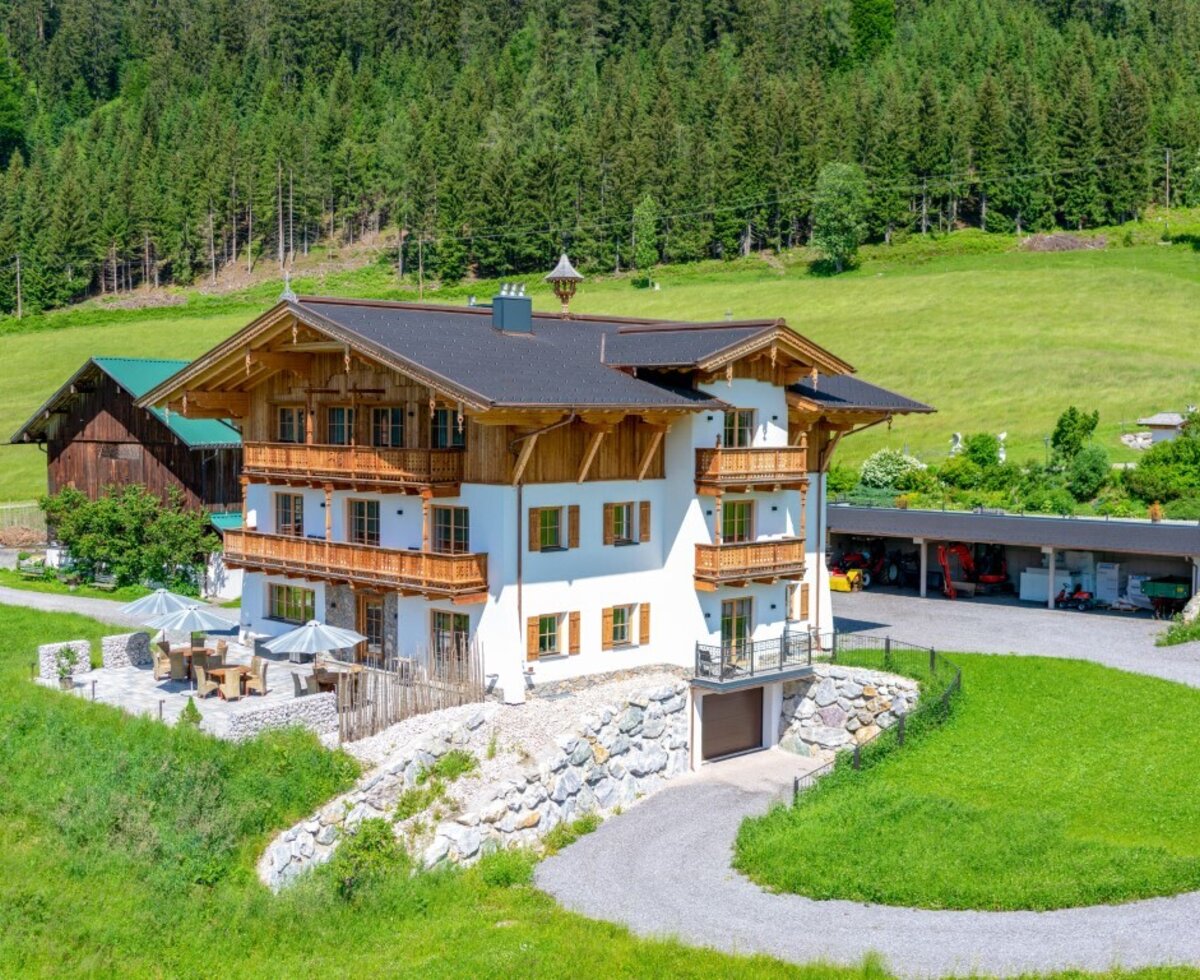 Der Bauernhof mit seinen Holzbalkonen und der Außenterrasse liegt in einer grünen Berglandschaft.