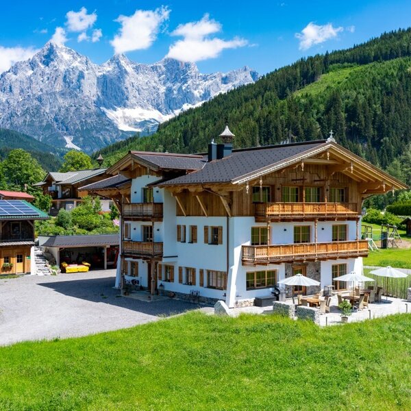 Der Bauernhof mit traditioneller Holzfassade, mehreren Balkonen und einer Terrasse mit Sitzgelegenheiten, umgeben von grünen Wiesen und Bergblick.