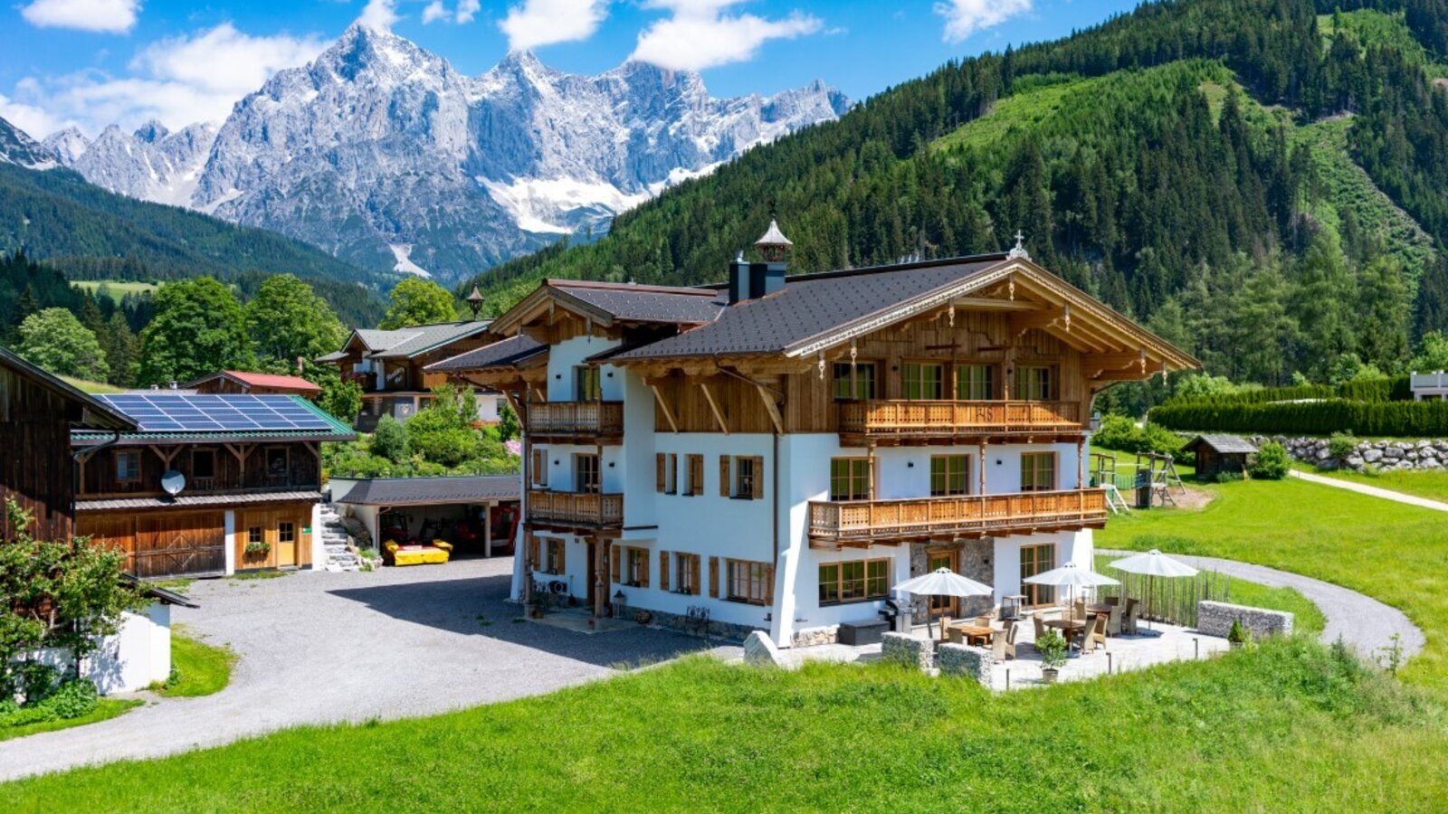 Der Bauernhof mit traditioneller Holzfassade, mehreren Balkonen und einer Terrasse mit Sitzgelegenheiten, umgeben von grünen Wiesen und Bergblick.