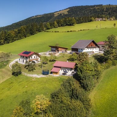 Der Hof mit seinen Gebäuden, inklusive eines mit Solarpaneelen, auf einer grünen Anhöhe umgeben von Wiesen und Wäldern.