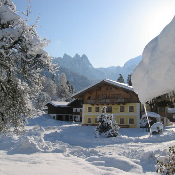 Der Bauernhof in einer verschneiten Winterlandschaft mit gelber Fassade, Holzbalkonen und Bergblick.