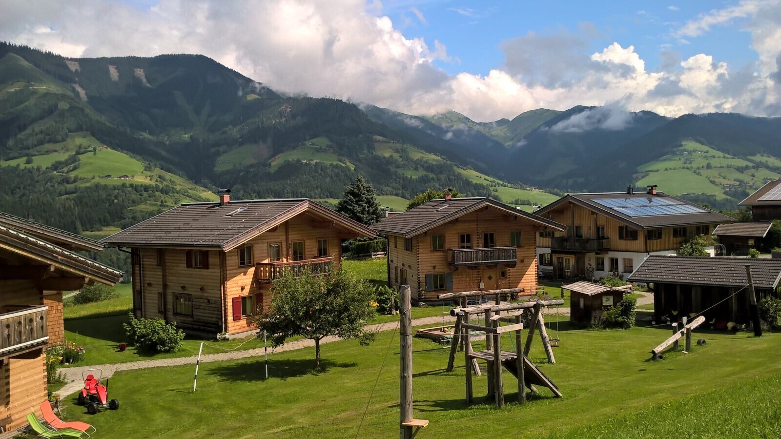 Der Bauernhof bietet mehrere Holzhäuser auf einer grünen Wiese mit Kinderspielplatz und Bergblick.