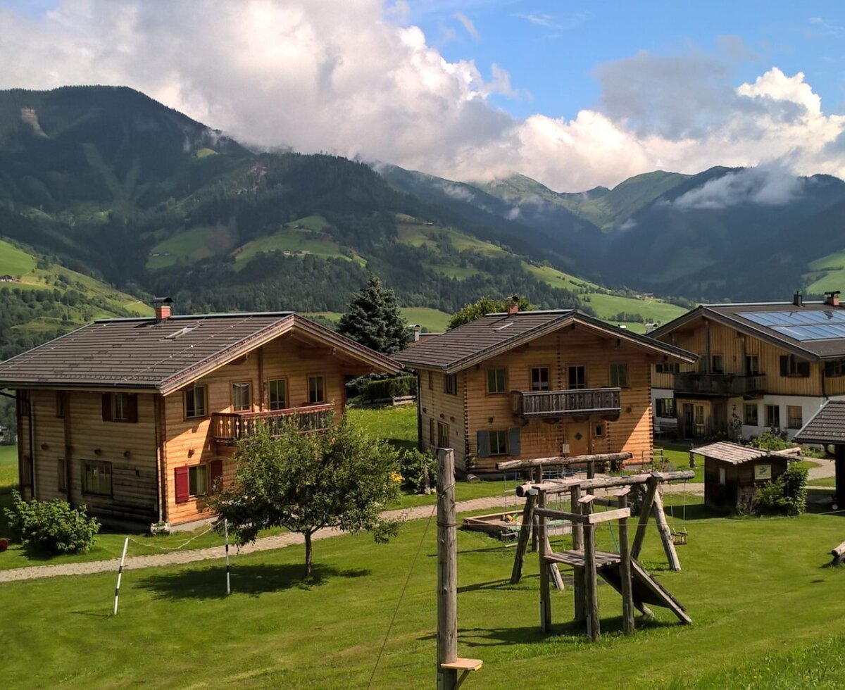 Der Bauernhof bietet mehrere Holzhäuser auf einer grünen Wiese mit Kinderspielplatz und Bergblick.