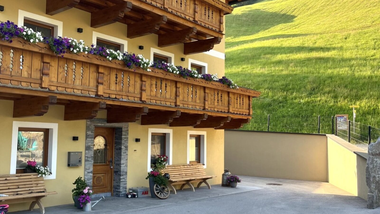 Der Bauernhof von außen mit traditionellen Holzbalkonen und Blumenkästen, einer gepflasterten Zufahrt und einem Blick auf die grünen Hügel.