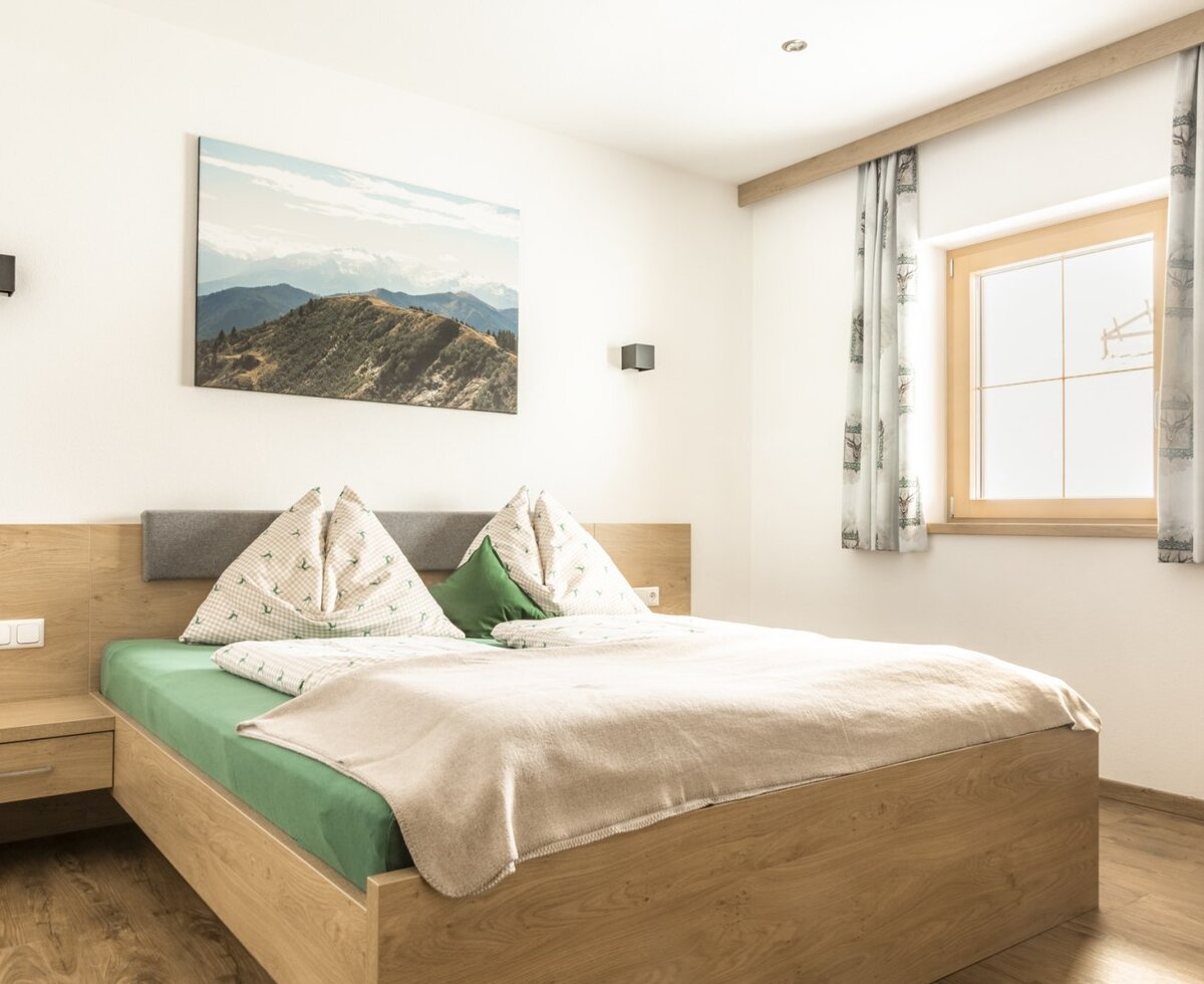 Doppelzimmer mit Holzbett, Nachttisch und Fenster mit Vorhängen.