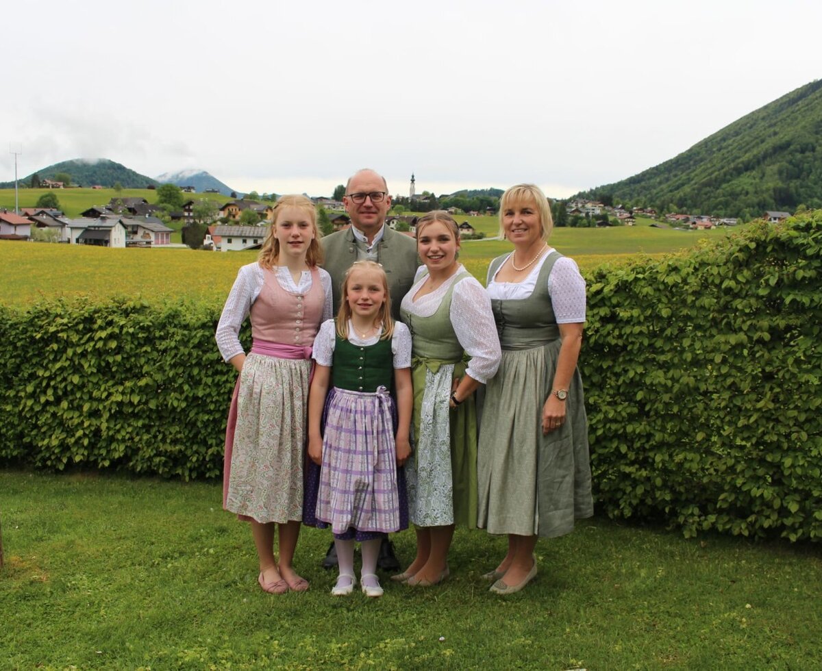 Die herzliche Gastfamilie des Bauernhofs in traditioneller Tracht vor einer malerischen, grünen Bergkulisse.