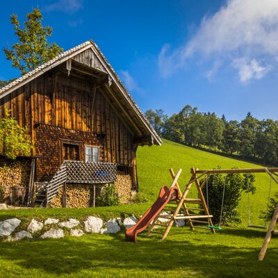 Der Bauernhof mit Kinderspielplatz im Garten, ausgestattet mit Rutsche und Schaukeln, vor einer grünen Hügellandschaft.