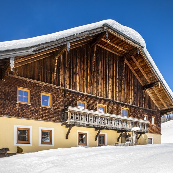 Der Bauernhof in winterlicher Umgebung, mit Schnee bedeckt, traditioneller Holzfassade, Balkon und einem Skilift im Hintergrund.