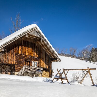 Der Bauernhof im Winter mit schneebedecktem Dach, Holzfassade, gestapeltem Holz und einer Holzschaukel.