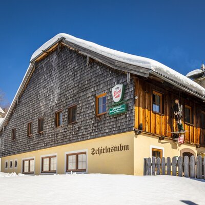 Der Bauernhof im Winter, mit einer Fassade aus Holzschindeln und gelbem Putz, sowie dem Gasthaus "Scharla Stubn" und verschneiter Umgebung.