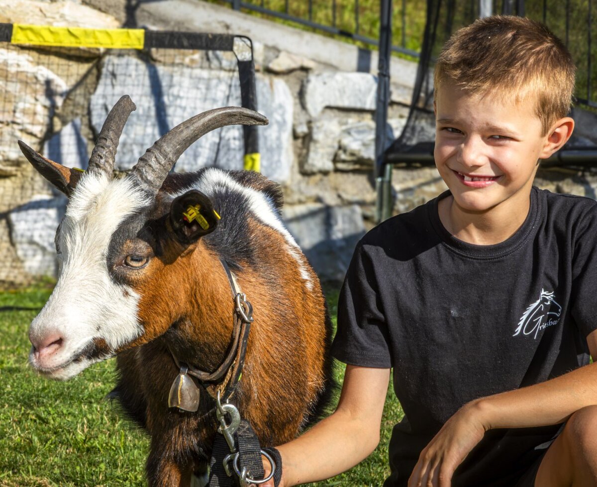Ein Junge lächelt neben einer zahmen Ziege auf der Wiese des familienfreundlichen Bauernhofs.