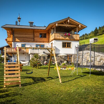 Der einladende Garten des Bauernhofs mit einer Schaukel, einem Trampolin und einer gepflegten Rasenfläche vor der malerischen Bergkulisse.