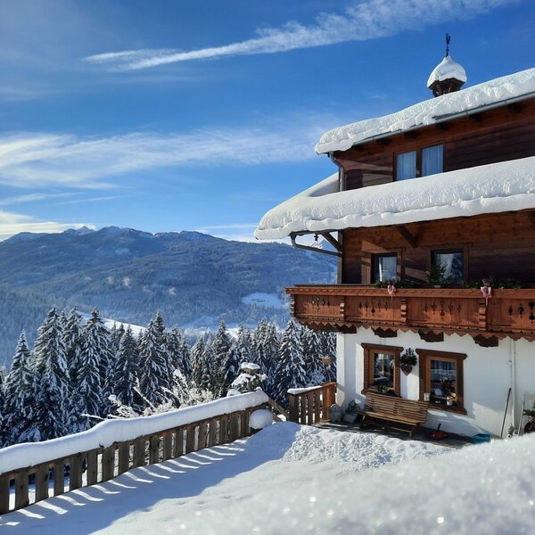Blick vom verschneiten Bauernhof auf die winterliche Berglandschaft und die schneebedeckten Wälder.