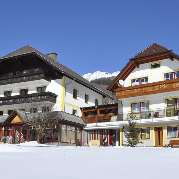 Außenansicht des Bauernhofs im Winter, mit schneebedecktem Vorplatz, traditionellen Balkonen und Bergen im Hintergrund.