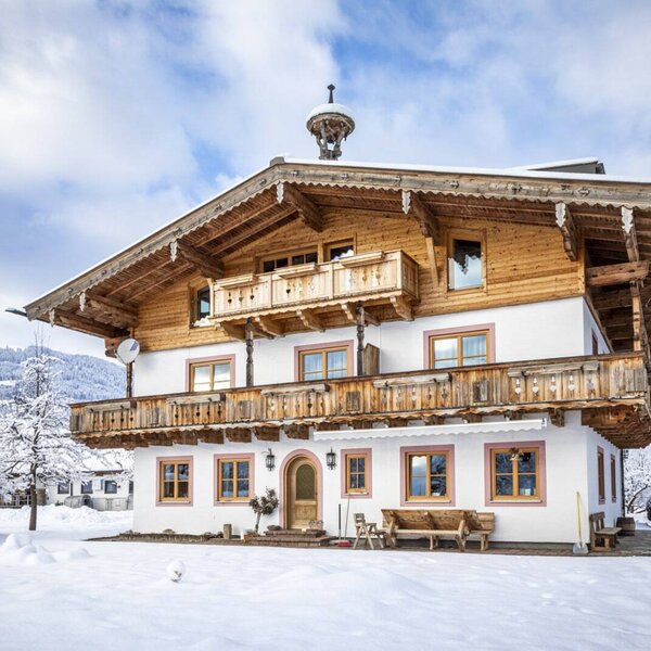 Der Bauernhof mit traditioneller Holzfassade, mehreren Balkonen und verschneitem Außenbereich vor winterlicher Berglandschaft.