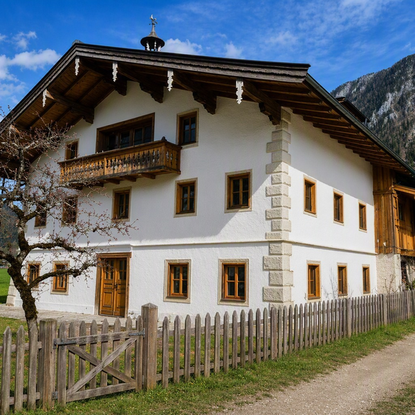Ein traditioneller Bauernhof mit einem charmanten Holzbalkon und einem gepflegten Holzzaun vor einer malerischen Bergkulisse.