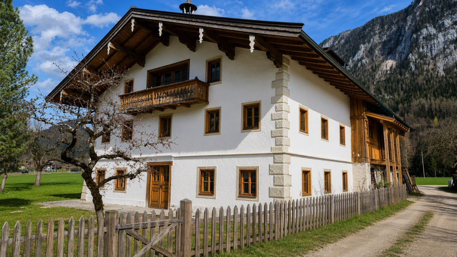 Ein traditioneller Bauernhof mit einem charmanten Holzbalkon und einem gepflegten Holzzaun vor einer malerischen Bergkulisse.