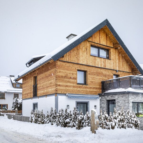 Das Äußere des Bauernhofs im Winter, mit verschneitem Dach, Holzfassade und einem Steinanbau mit Balkon.