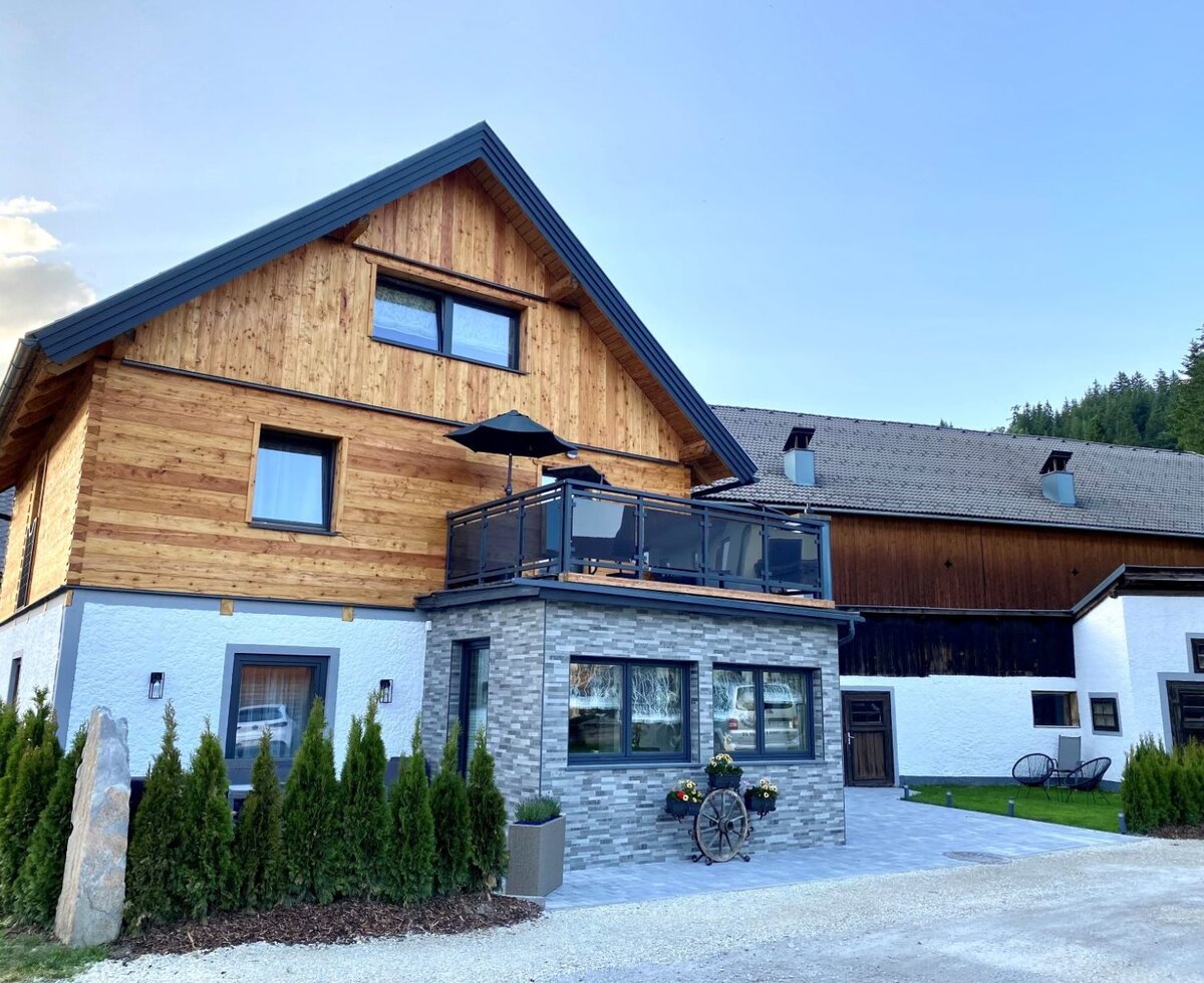 Die Außenansicht des Bauernhofs mit modernisierter Holzfassade, einem Balkon mit Sitzgelegenheiten und Sonnenschirm sowie dem angrenzenden Wirtschaftsgebäude.