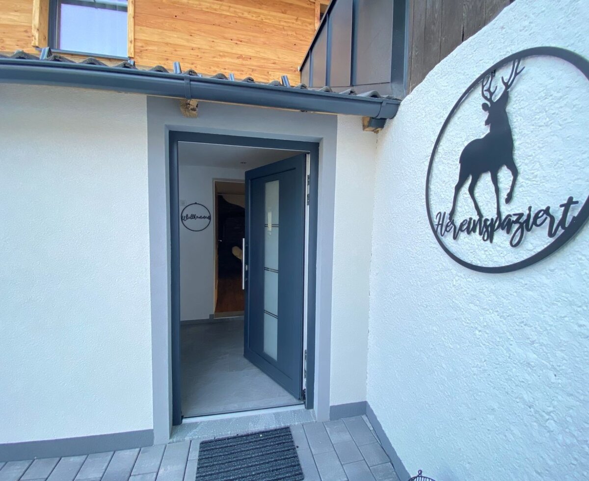 Der Eingangsbereich des Bauernhofs mit offener Tür, einem "Hereinspaziert"-Schild mit Hirsch-Silhouette und einem "Willkommen"-Schild innen.