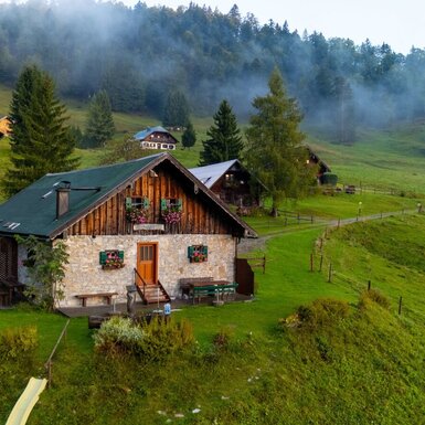 Der Hof, gebaut aus Stein und Holz, liegt auf einer grünen Wiese mit Hügel- und Waldblick und bietet eine Rutsche sowie Sitzgelegenheiten im Außenbereich.