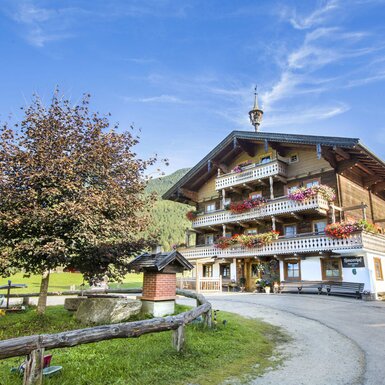 Der Bauernhof mit traditioneller Holzfassade, mehreren Etagen und blumengeschmückten Balkonen, umgeben von grünen Wiesen und Wäldern.