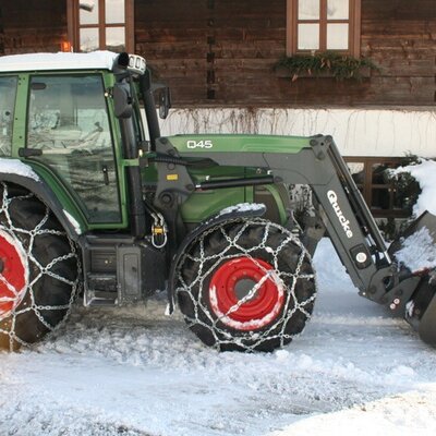 Ein grüner Traktor mit Schneeketten vor dem rustikalen Holzgebäude des Bauernhofs im Winter.