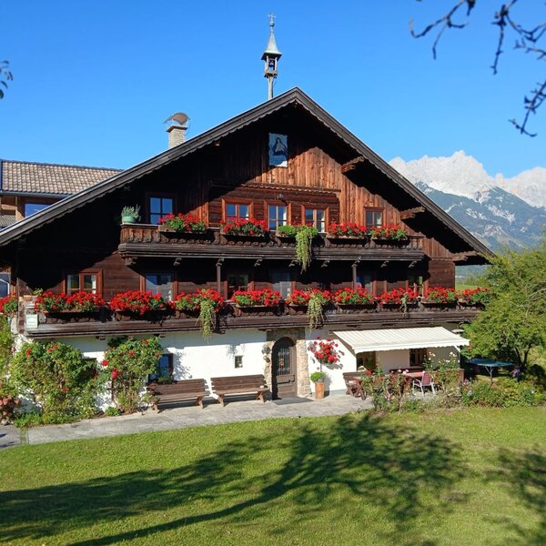 Der einladende Bauernhof Hinterburghof mit blumengeschmückten Holzbalkonen und einer sonnigen Liegewiese vor der malerischen Bergkulisse.