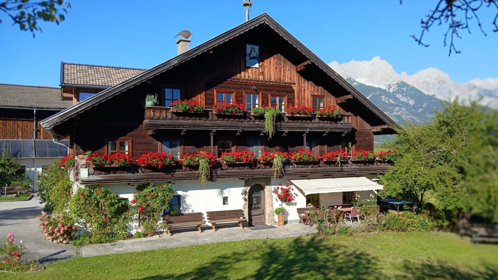 Der einladende Bauernhof Hinterburghof mit blumengeschmückten Holzbalkonen und einer sonnigen Liegewiese vor der malerischen Bergkulisse.