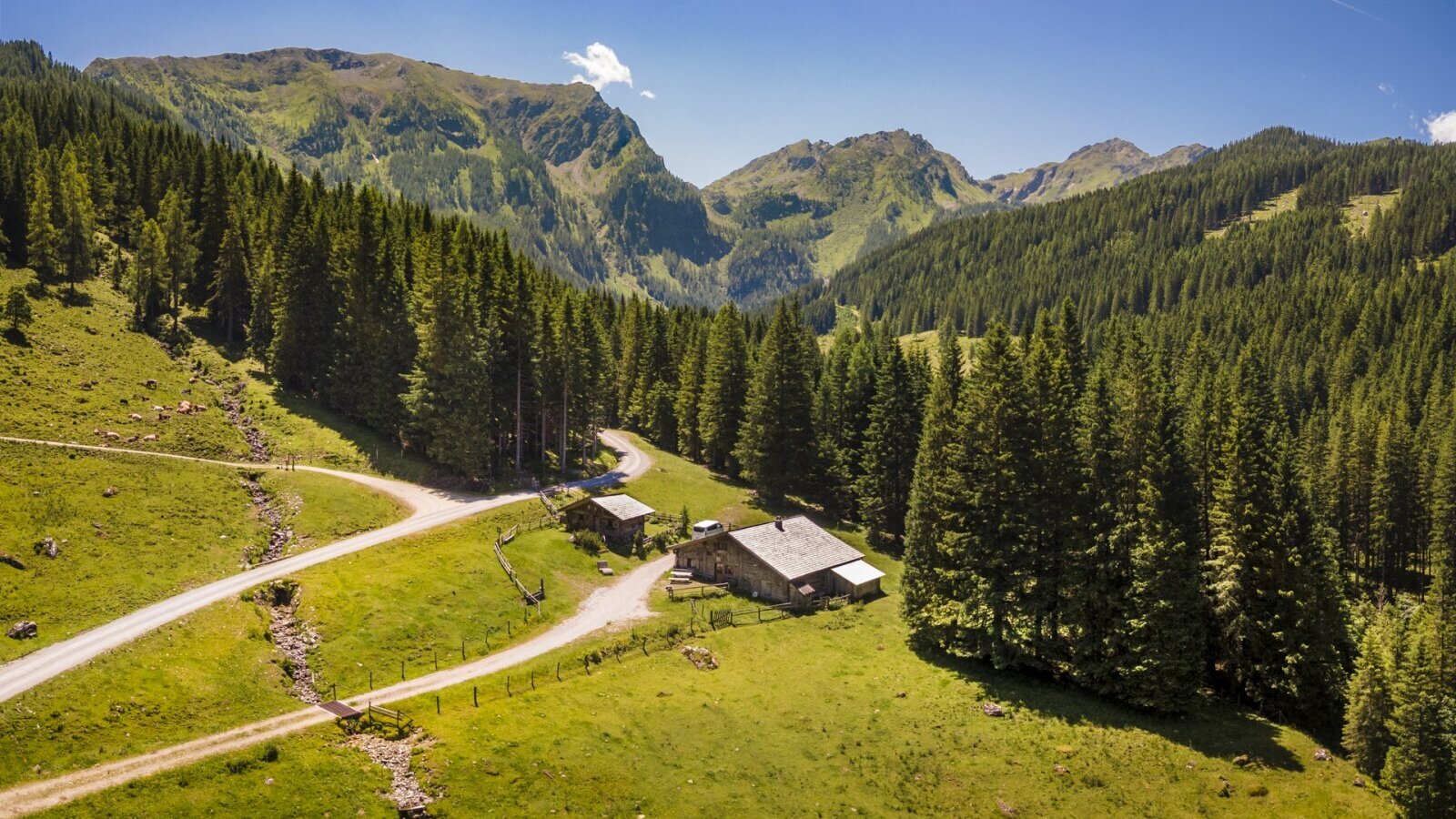Der Hof liegt in einem Bergtal, umgeben von Nadelwäldern und grünen Wiesen, mit einer Zufahrtsstraße.
