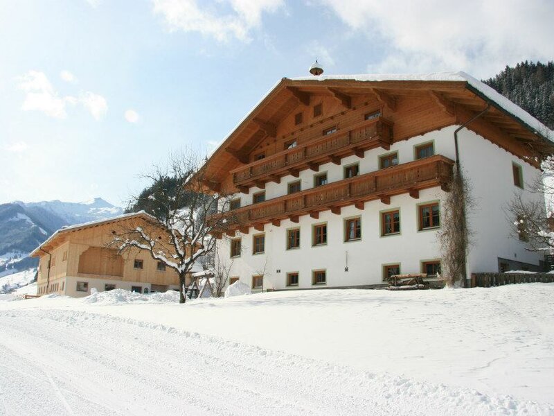 Der Bauernhof mit Nebengebäude im Winter, mit weißer Fassade, Holzbalkonen und Holzgiebel vor einer verschneiten Bergkulisse.