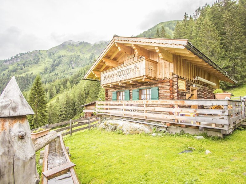 Der traditionelle Hof aus Holz mit Balkon und grünen Fensterläden liegt auf einer Grünfläche vor einem Berghang und verfügt über einen Holzwassertrog im Vordergrund.